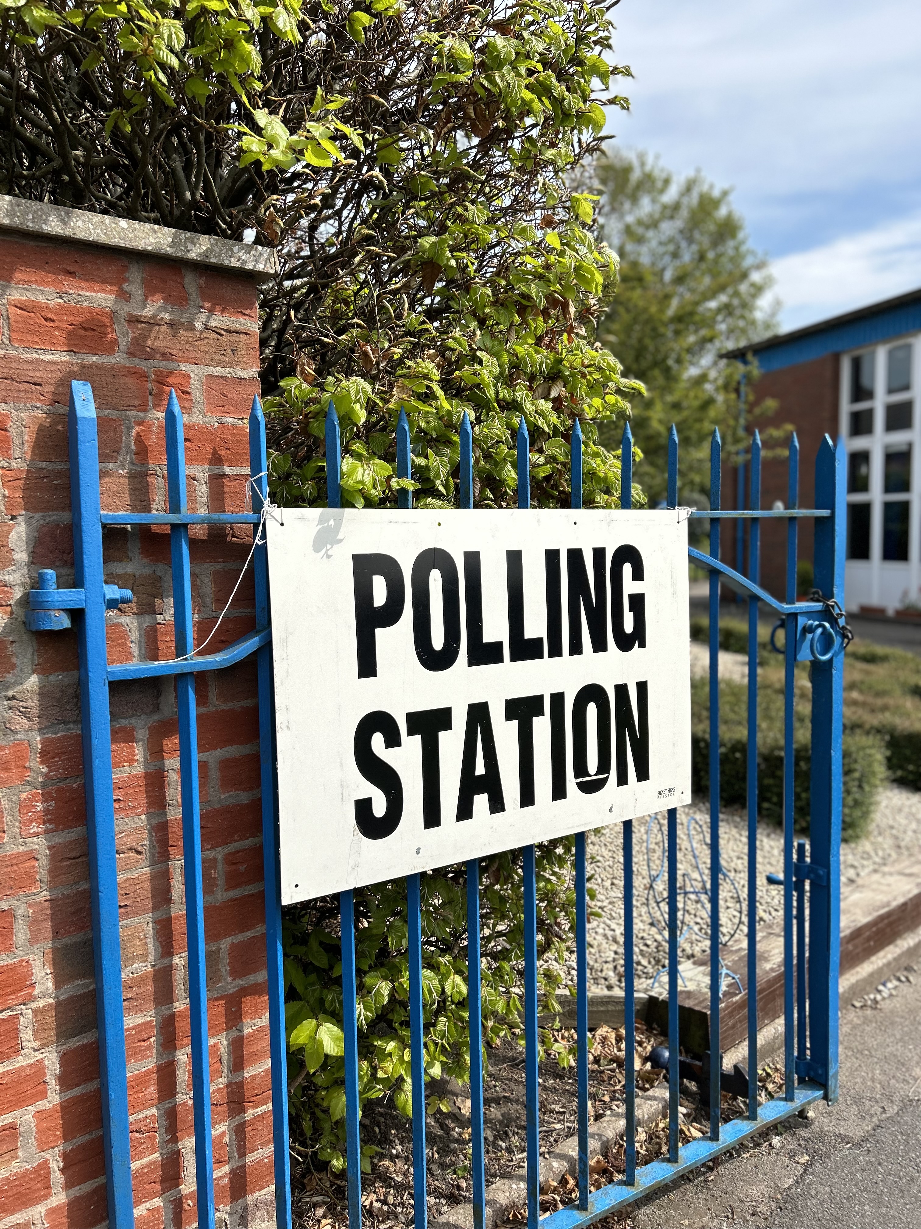 'polling station' sign on a blue fence next to a brick wall and a tree