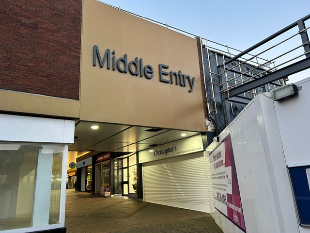 entrance to shops showing a gold bridge above alley to shops and hoarding to the right