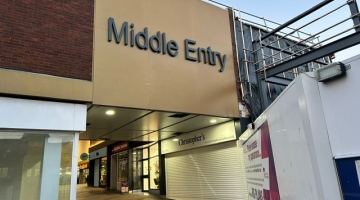 entrance to shops showing a gold bridge above alley to shops and hoarding to the right